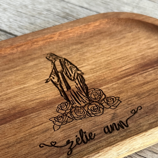 Oval wooden tray with image of Mary with roses at her feet. Under image is an engraved name.